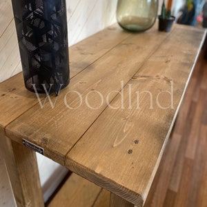 May include: A rustic wooden console table with a natural finish. The table features a rectangular top made of wooden planks and sturdy legs. A black metal lantern and a small metal tag with the word "Woodind" are visible on the table. The table is suitable for entryways or living rooms.