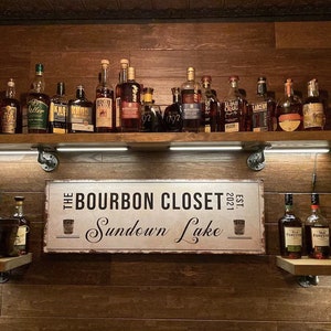 May include: A metal sign with the text "The Bourbon Closet Sundown Lake Est. 2021" and a small illustration of a glass of bourbon. The sign is hanging on a wooden wall with shelves of various bottles of bourbon.