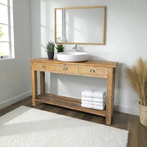 Peut inclure: Meuble-lavabo en bois rustique avec vasque blanche, trois tiroirs et une étagère inférieure. Un miroir rectangulaire est suspendu au-dessus de la vanité. Des serviettes blanches sont empilées sur l'étagère. Deux petites plantes en pot sont posées sur le dessus.