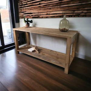 May include: A rustic, wooden console table with a lower shelf. The table is made of light brown wood and has a rectangular shape. A glass vase and a small potted plant sit on top. The table is 60 inches long, 15 inches wide, and 30 inches high.