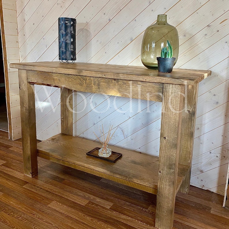 May include: A rustic wooden console table with a lower shelf. The table is made of light brown wood and has a natural finish. Decorative items on the table include a blue metal lantern, a green glass vase, and a small potted plant. The table is 60 inches long.