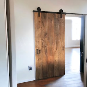 May include: A rustic wooden sliding barn door with black hardware and a metal handle. The door is open, revealing a room with a white wall and a wooden floor.