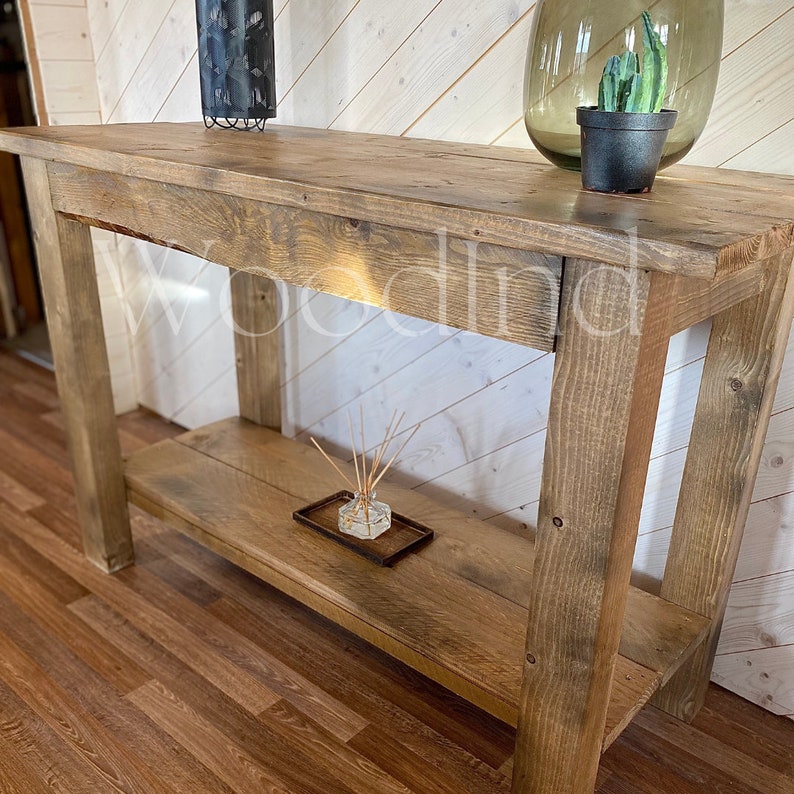 May include: A rustic, wooden console table with a lower shelf. The table is made of light brown, weathered wood. A small black pot with a green plant and a decorative glass vase are on the top surface. A glass diffuser sits on the lower shelf.