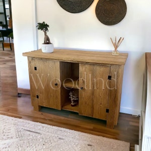 Könnte beinhalten: Ein rustikaler Holzschrank mit zwei Türen und einem Regal. Der Schrank hat eine natürliche Holzoptik und schwarze Metallbeschläge. Der Schrank steht auf einem Holzboden.
