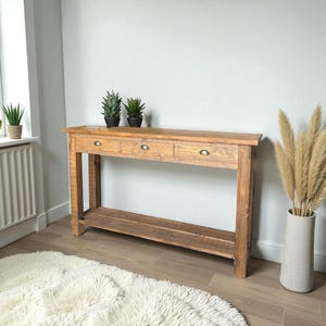 Puede incluir: Una consola de madera marrón claro con tres cajones y un estante inferior. La mesa tiene una parte superior rectangular y está sostenida por cuatro patas robustas. Los cajones tienen pequeños tiradores redondos de metal. Dos pequeñas plantas en maceta están encima de la mesa.