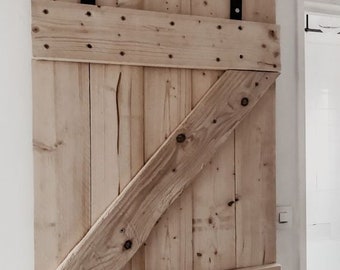 May include: A rustic wooden barn door with a black metal sliding track. The door is constructed from light-colored wooden planks, with a diagonal brace and horizontal supports. The hardware is black, and the door is mounted on a white wall.