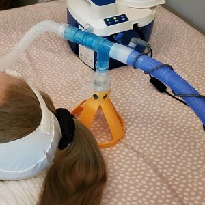 May include: A young person with long brown hair is lying on a bed with a white medical mask and a blue and white medical device connected to a clear tube. The device is on a white and blue base with a yellow stand. The bed has a pink and white patterned sheet.