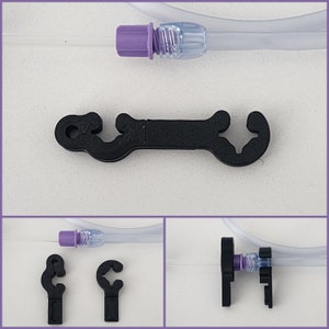 May include: A black plastic clamp with a hook on each end. The clamp is designed to hold a medical tube in place. There are two additional clamps in the image, one on the left and one on the right.