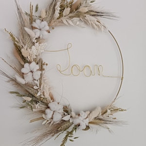 Op de afbeelding: Een gedroogde bloemenkrans met een gouden metalen ring en het woord "kamer" geschreven in gouden draad. De krans is gemaakt van gedroogde grassen, katoenen bollen en andere natuurlijke elementen.