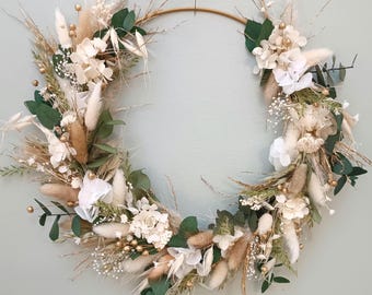 May include: A circular wreath featuring a gold metal ring base, adorned with dried flowers and greenery. The arrangement includes white hydrangeas, fluffy beige grasses, and dark green eucalyptus leaves, creating a natural and elegant aesthetic.