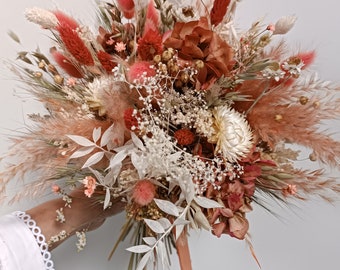 Può includere: Un bouquet di fiori secchi con un mix di fiori bianchi, rosa e marroni. Il bouquet è legato con un nastro rosa.