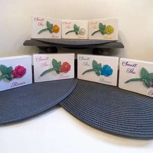 May include: Six wooden signs with the text "Smell the Roses" and a 3D rose in different colors: pink, orange, white, yellow, blue, and purple.