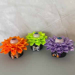 May include: Three decorative circular ornaments featuring layered fabric petals in bright orange, neon green, and light purple. Each ornament is topped with a central white sphere painted to resemble a human eyeball with a blue iris and red veins.