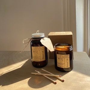 May include: Two amber glass candle jars with brown labels and the text "Patchouli, Lavender & Pink Grapefruit" and "Extra Long Safety Matches" on them. The candles are on a wooden surface with a box and matches.