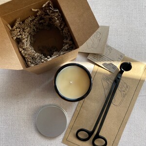 May include: A gift set for candle enthusiasts, containing a black candle in a glass jar, a small silver tin, and a black candle snuffer with a wooden handle. The set is presented in a brown cardboard box filled with shredded paper.