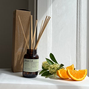 May include: A brown glass bottle diffuser with a brown label that reads "Boucl&eacute; Essential Oil Diffuser Ylang Ylang & Mandarin Zest" and a set of wooden sticks. The bottle is sitting on a white surface with a small bouquet of white flowers and orange slices.