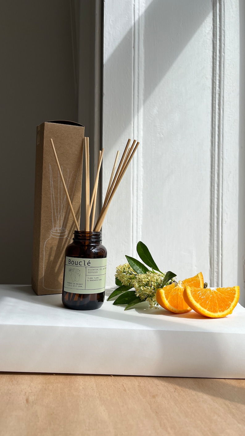 May include: A brown glass bottle diffuser with a brown label that reads "Boucl&eacute; Essential Oil Diffuser Ylang Ylang & Mandarin Zest" and a set of wooden sticks. The bottle is sitting on a white surface with a small bouquet of white flowers and orange slices.