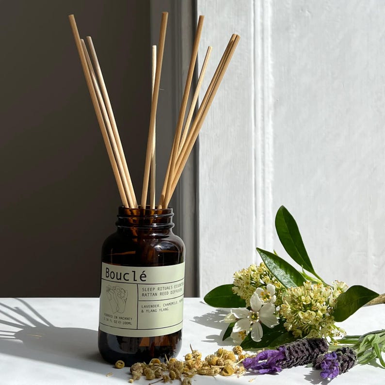 May include: A brown glass bottle with a label that reads "Boucl&eacute; Sleep Rituals Essential Oil Rattan Reed Diffuser Lavender, Chamomile & Ylang Ylang" and a set of wooden sticks in the bottle. The bottle is sitting on a white surface with dried flowers and lavender.