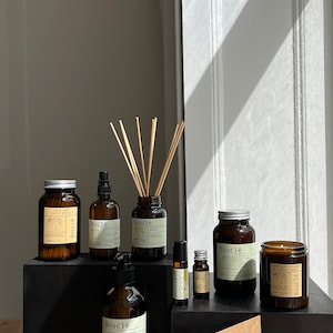 May include: A collection of brown glass bottles with labels that say "Boucl&eacute;" and contain various products, including essential oils, hand soap, candles, and a diffuser. The bottles are arranged on a black surface with a brown card with a black and white illustration of two hands.
