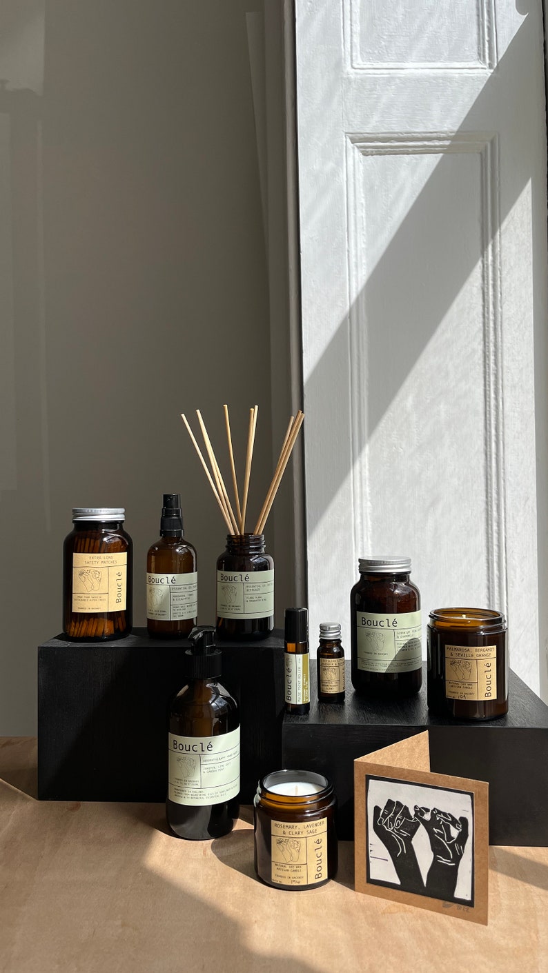 May include: A collection of brown glass bottles with labels that say "Boucl&eacute;" and contain various products, including essential oils, hand soap, candles, and a diffuser. The bottles are arranged on a black surface with a brown card with a black and white illustration of two hands.