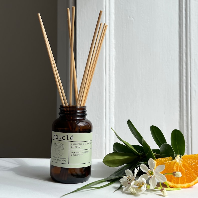 May include: A brown glass bottle diffuser with a label that reads "Boucl&eacute; Essential Oil Natural Diffuser Palmarosa, Bergamot & Eucalyptus" and wooden sticks sticking out of the top. The bottle is sitting on a white surface with a sprig of orange blossoms and an orange slice.