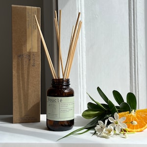 May include: A brown glass bottle with a brown label that reads "Boucl&eacute; Essential Oil Diffuser Palmarosa, Bergamot & Eucalyptus" and a brown cardboard box with the text "Boucl&eacute;" and "Palmarosa, Bergamot & Eucalyptus Pure Essential Oil Rattan Diffuser" on it. The bottle is filled with a clear liquid and has a set of wooden sticks in it. The bottle is sitting on a white surface.