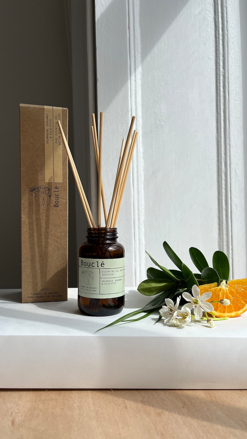 May include: A brown glass bottle with a brown label that reads "Boucl&eacute; Essential Oil Diffuser Palmarosa, Bergamot & Eucalyptus" and a brown cardboard box with the text "Boucl&eacute;" and "Palmarosa, Bergamot & Eucalyptus Pure Essential Oil Rattan Diffuser" on it. The bottle is filled with a clear liquid and has a set of wooden sticks in it. The bottle is sitting on a white surface.