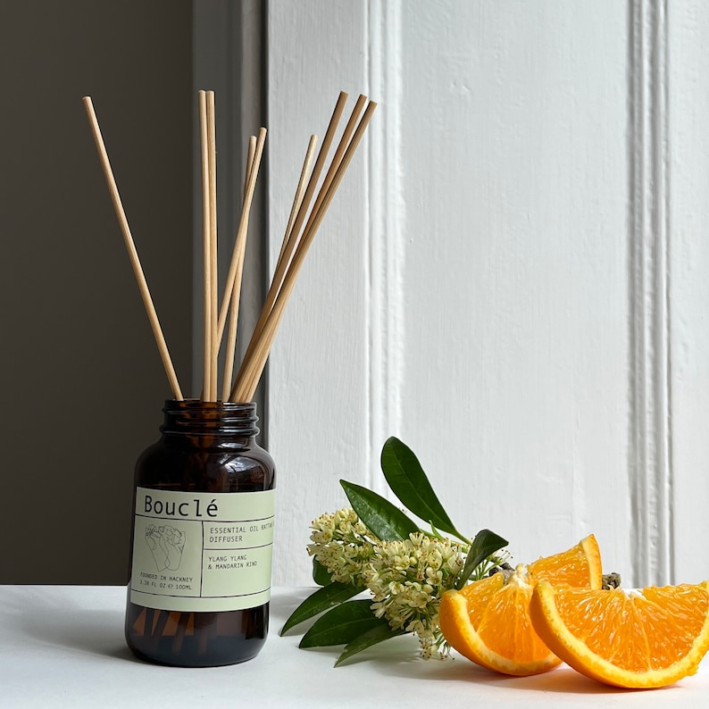 May include: A brown glass bottle diffuser with a label that reads "Boucl&eacute; Essential Oil Diffuser Ylang Ylang & Mandarin Zest" and a set of wooden sticks. The bottle is sitting on a white surface with a sprig of white flowers and orange slices.