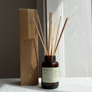 May include: A brown glass bottle with a brown label that reads "Boucl&eacute; Essential Oil Diffuser Petitgrain, Palmarosa & Saint-Benoit Rose 100ml Founded in Hackney Hand Poured in England" with wooden sticks in the bottle.