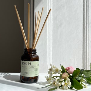 May include: A brown glass bottle diffuser with a label that reads "Boucl&eacute; Essential Oil Diffuser Petitgrain, Palmarosa & Saint-Benoit Rose" and a bouquet of white and pink flowers.