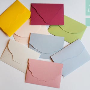 Può includere: Una collezione di buste colorate in varie tonalità, tra cui giallo, rosa e blu, sono disposte su una superficie bianca. Le buste hanno un esclusivo design con patta smerlata. Il testo nell'immagine indica 43 colori e 10 forme disponibili.
