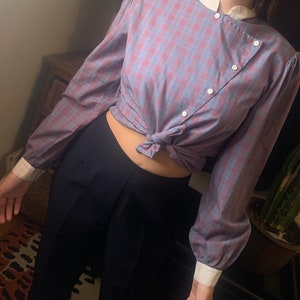 Pode incluir: Uma camisa de manga comprida xadrez azul e rosa com colarinho e punhos brancos. A camisa é amarrada na cintura e tem um único fecho de botão na frente.