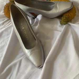 May include: A pair of silver high-heeled shoes with a square toe. The shoes have a silver metal accent on the toe and are labeled "MAR STUDIO" on the inside.