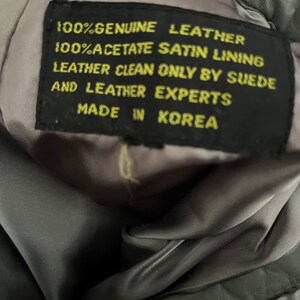 May include: Close-up of a black leather tag with yellow text that reads "100% Genuine Leather 100% Acetate Satin Lining Leather Clean Only By Suede And Leather Experts Made In Korea."