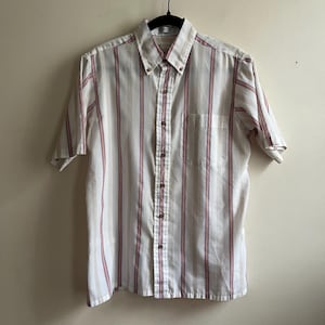 May include: Short-sleeved shirt with vertical stripes in white, beige, and red. The shirt features a button-down collar and a chest pocket. It is hanging on a black hanger.