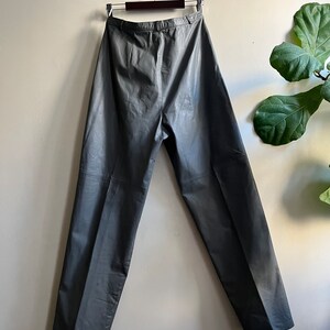 May include: Gray leather pants hanging on a wooden hanger against a white wall. The pants have a high waist and a straight leg.