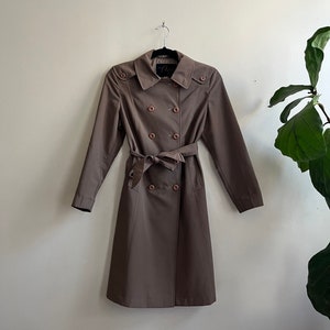 May include: A brown double-breasted trench coat with a belt and button closures. The coat is hanging on a hanger against a white wall.