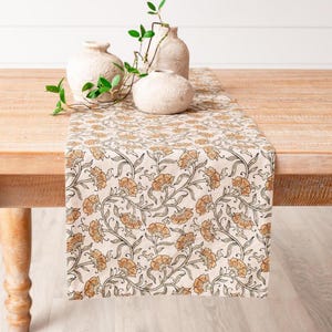 Op de afbeelding: Een beige tafelloper met een herhalend bloemmotief in tinten oranje en bruin. De loper ligt op een houten tafel en er zijn drie kleine, getextureerde vazen en groen op geplaatst.