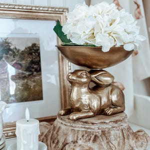 May include: A gold-colored bunny figurine atop a tree stump base, supporting a gold bowl filled with white flowers. A framed picture and a candle on a stack of saucers are in the background. The overall theme is Easter decor.