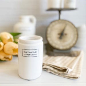 Może przedstawiać: Biały ceramiczny słoik z napisem "WHITE COTTAGE STONEWARE CO." w czarnym prostokącie. Słoik jest cylindryczny z lekko rozszerzoną krawędzią. W tle znajduje się zabytkowa waga i beżowa kraciasta ściereczka.
