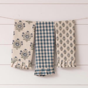 Op de afbeelding: Drie decoratieve keukendoeken hangen aan een waslijn. Twee handdoeken hebben een bloemenpatroon in blauw en beige, met een gerimpelde rand. De middelste handdoek heeft een blauw en wit geruit patroon, ook met een gerimpelde rand. De handdoeken hangen tegen een witte houten achtergrond.