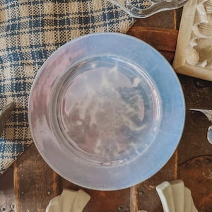 May include: A circular glass plate featuring an iridescent finish that reflects light with soft blue and purple hues. The plate has a smooth, flat center and a slightly raised rim, showcasing a reflective surface that creates a subtle, shimmering effect.