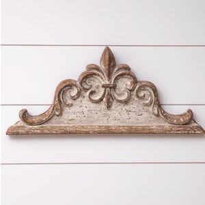 May include: A distressed, wooden shelf with a fleur-de-lis design. The shelf is painted in a cream color with brown accents and features ornate scrollwork. The shelf is mounted on a white, horizontal-slatted wall.