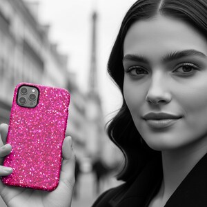 May include: A vibrant hot pink smartphone case with a dense, sparkling glitter finish. The case features a rectangular cutout for a triple-lens camera system on the upper left corner, showcasing a textured, reflective surface that catches light.