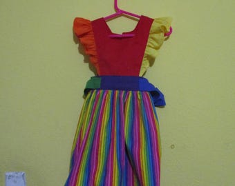 Puede incluir: Un vestido infantil colorido con una falda a rayas arcoíris y un corpiño rojo. El vestido tiene mangas con volantes amarillas y naranjas, y un cinturón azul. Está colgado de una percha rosa.
