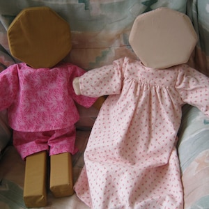 May include: Two handmade fabric dolls with pink floral print dresses. The dolls have brown and beige heads and are sitting on a patterned fabric surface.