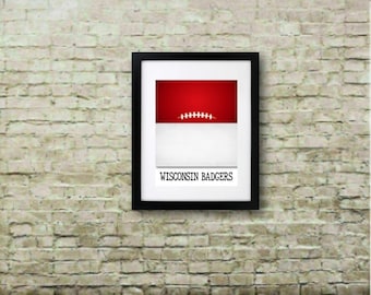 May include: Framed art print with a red and white design and a football graphic. The artwork includes the text "WISCONSIN BADGERS" in a black frame, hanging on a brick wall. The floor is made of wooden planks.