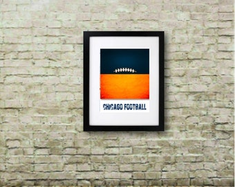 May include: Framed art print with a minimalist football design against a blue and orange backdrop. The text "CHICAGO FOOTBALL" is at the bottom. The artwork is in a black frame and hangs on a brick wall. The room has a wooden floor.