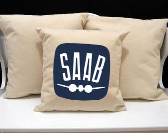 May include: Three beige square cushions. The central cushion displays a navy blue square with the word "SAAB" in white, and a white graphic below. The cushions are arranged on a white surface.