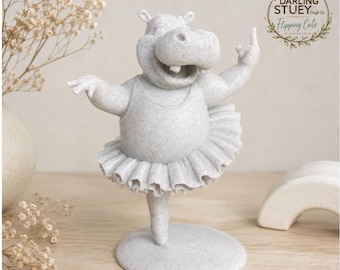 May include: A light grey speckled hippo figurine wearing a tutu and leotard, posed in a ballet stance on a circular base. The hippo has an open mouth and one arm raised. A logo in the top right corner reads DARLING STUEY Craft Co. Flipping Cute.